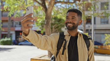 Sakallı, gülümseyen genç bir adam güneşli bir şehir parkında sırt çantası takarak selfie çekiyor..