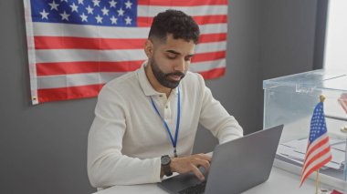 Amerikan bayrağı ve seçim sandığı olan bir odada sakallı İspanyol bir adam dizüstü bilgisayarla çalışıyor..