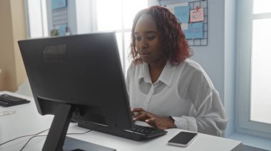 Ofisteki bilgisayarda çalışan genç Afro-Amerikalı bir kadın masada akıllı bir telefonla modern ve üretken bir iş ortamının havasını yakalıyor..