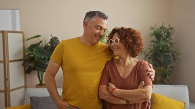 Orta yaşlı çift rahat oturma odalarında sıcacık kucaklaşıyorlar, iç mekanlarında bitkiler ve arka planda modern dekorlar var.