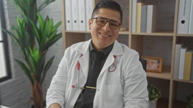 A smiling hispanic male doctor in a white coat sits confidently in a hospital office, portraying professionalism and friendliness. clipart