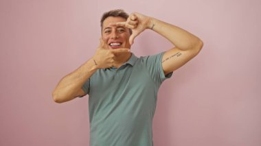 Genç bir adam, çekici ve gülümseyen, pembe bir arka planda soyutlanmış, ellerini yaratıcı bir şekilde poz verip çerçeveliyor..