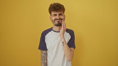 A young hispanic man with tattoos winces in pain holding his cheek against a yellow background. clipart