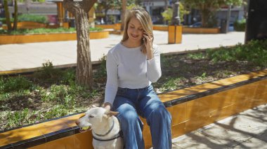 Gülümseyen genç bir kadın neşeli köpeğiyle oturup akıllı telefonuyla konuşurken güneşli bir şehir parkından zevk alıyor..