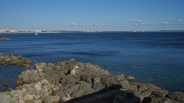 Puglia, İtalya 'da Gelibolu' nun sakin kıyıları kayalık kıyıları, berrak mavi suları ve parlak gökyüzü altında uzak bir yelkenli sergiliyor..