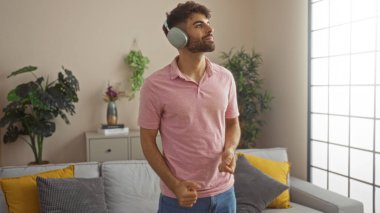 Modern bir oturma odasında kulaklık takan sakallı genç bir adam, pembe gömlek ve kot pantolon giyerken müzikten zevk alıyor, bitkiler ve hoş bir dekorla çevrili..