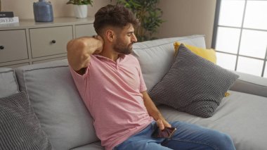 Oturma odasındaki koltukta oturan sakallı genç adam boynunu uzatırken telefonu tutuyor ve modern dekorlu rahat bir iç mekan ortamı sergiliyor.