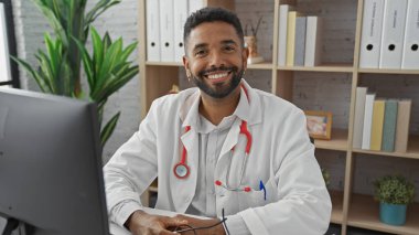 Steteskoplu beyaz önlüklü gülümseyen Afro-Amerikan erkek doktor hastane ofisindeki bir masada oturuyor.