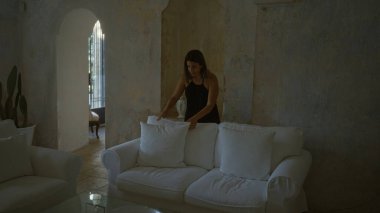 Genç bir İspanyol kadın, tipik bir İtalyan masseryasında beyaz bir kanepeye yastık yerleştiriyor ve rahat bir ev ortamı yaratıyor..