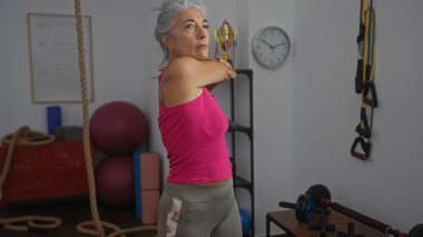 Spor salonunda esneyen gri saçlı olgun bir kadın. Spor salonunun etrafı spor aletleriyle çevrili.