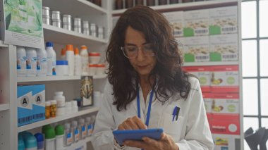Bir eczanede çalışan İspanyol kadın eczacı elinde arka planda raflar dolusu ürünler olan bir tablet tutuyor..