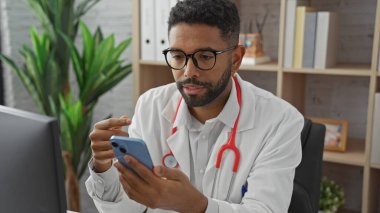 Sakallı Afrikalı Amerikalı erkek doktor klinikte akıllı telefon kullanıyor.