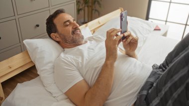 Yatakta uzanmış İspanyol bir adam, yatak odası ortamında akıllı telefon kullanıyor, rahat ve modern bir yaşam tarzı sergiliyor..