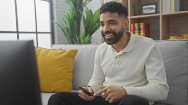 Yakışıklı, sakallı bir İspanyol, elinde kumandayla oturmuş gülümsüyor..