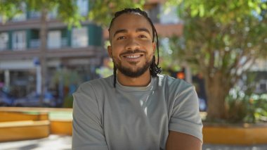 Saçları örgülü yakışıklı genç adam güneşli bir günde şehir parkında kendinden emin bir şekilde gülümsüyor..