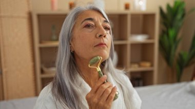 Mature woman with grey hair using a facial roller in a wellness center with spa-like interior clipart