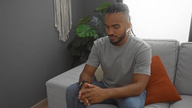 Saçları örgülü genç bir adam rahat bir oturma odasında, rahat kıyafetler giymiş ve derin düşüncelere dalmış..