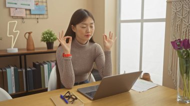 Genç Asyalı bir kadın, ev dekorasyonu ve kitaplarla çevrili rahat dairesinde farkındalık pratiği yapıyor..