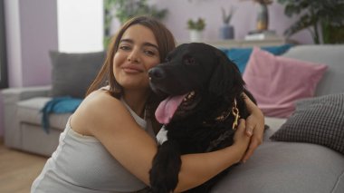 Siyah labrador köpeğini modern dekor ve iç mekan bitkileriyle sıcak bir oturma odasında kucaklayan genç İspanyol kadın, sıcaklık ve arkadaşlık saçıyor..