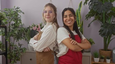İki kadın, kollarını kavuşturmuş, saksı bitkilerinin arasında yan yana duruyorlar, bir çiçekçi dükkanında takım çalışması yapıyorlar..