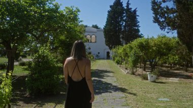 Siyah elbiseli genç İspanyol kadın Puglia, İtalya 'da güzel bir villaya doğru yürüyor. Etrafı yemyeşil ve açık hava bahçeleriyle çevrili..