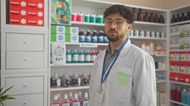 Sakallı genç profesyonel bir adam eczanede duruyor. Etrafı ürünlerle dolu raflarla çevrili..
