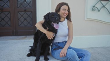 Güzel, genç, İspanyol bir kadın labrador köpeğiyle bir sokak ortasında oturuyor, neşeyle gülümsüyor..