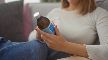 Bir kadın kanepede oturur, sıcak bir evde şurup şişesini inceler, sağlık ve ev hayatını yansıtır..