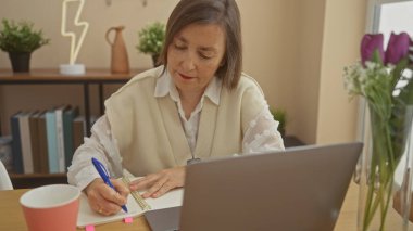 Evde dizüstü bilgisayarı, bitkileri ve dekoratif lambasıyla notlar yazan olgun bir kadın.