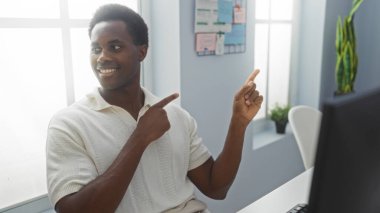 Genç adam gülümsüyor ve parlak, modern bir ofise işaret ediyor. Yönlendirme ya da fikir öneriyor. Profesyonel bir iş yeri ortamında görüntüleniyor.