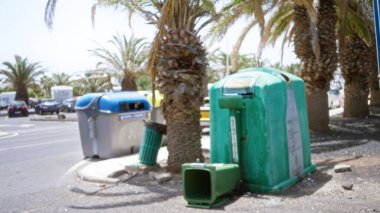 Arka planda bulanık palmiye ağaçları bulunan güneşli bir kentsel alanda, geri dönüşüm kutularını ve yerdeki çöpleri renklendiren çözülmüş atık konteynırları.