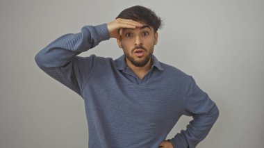 Sakallı, eli alnında, beyaz bir arka planda duran, şaşırmış genç bir adam..