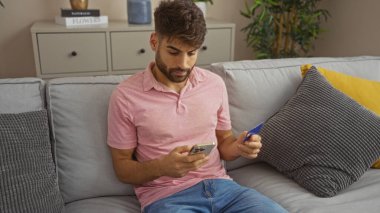 Genç, İspanyol, oturma odasında gri bir koltukta oturan, elinde kredi kartıyla cep telefonu kullanan, evdeki teknolojik etkileşime ve rahatlığa odaklanmış bir adam..