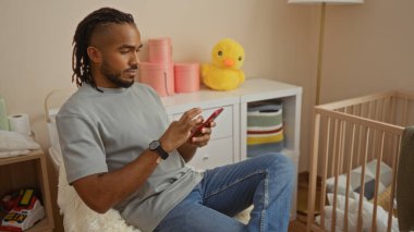 Yatak odasında oturan genç adam bebek beşiği ve oyuncaklarıyla ev dekorasyonunda telefon kullanıyor.