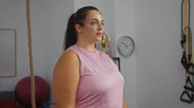 Artı, arka planda spor salonunda duran pembe bluzlu kadın.