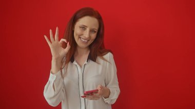 Beyaz bluzlu gülümseyen kızıl saçlı kadın kırmızı arka planda telefonla iyi bir hareket yapıyor.