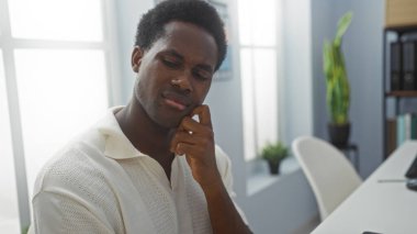 Modern ofis ortamında derin düşünen yakışıklı bir genç. Kapalı mekanları ve geniş pencereleri olan parlak doğal bir ışığı var.