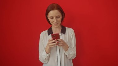 Kırmızı arka planda izole edilmiş akıllı telefon kullanan beyaz tişörtlü kızıl saçlı kadın.