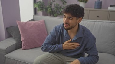 Gri bir kanepede otururken göğsünde ağrı hisseden sakallı yakışıklı genç bir adam..