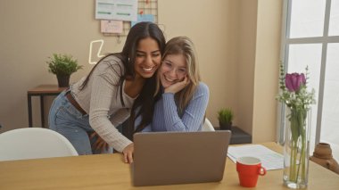 Modern bir oturma odasında dizüstü bilgisayar kullanan iki kadın neşeli bir anı paylaşıyor.