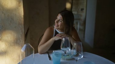 Genç, güzel bir İspanyol kadın İtalyan restoranında eğleniyor. Etrafında sofra takımı ve gözlüklerle pencereden dışarı bakıp gülümsüyor..