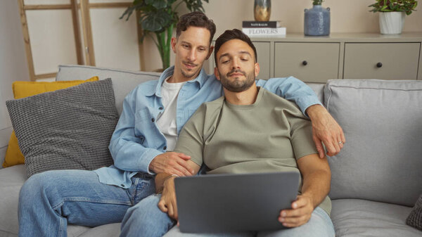 Двое мужчин в любящих отношениях расслабляются вместе дома на удобном диване, подчеркивая динамику гей-пары в уютном помещении.