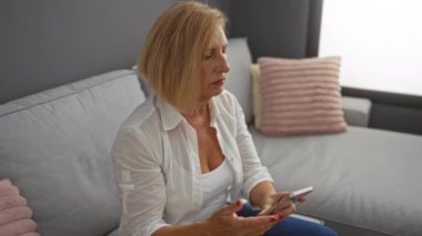 Modern bir oturma odasındaki koltukta oturan bir kadın akıllı telefon kullanıyor. Düşünceli ve düşünceli görünüyor. Kısa sarı saçlı ve beyaz gömlekli.