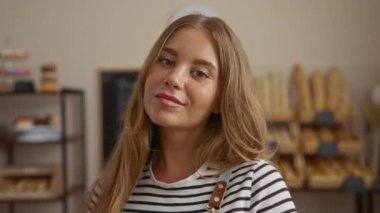 Bir fırında gülümseyen genç sarışın kadın arka planda ekmek raflarıyla, samimi ve davetkar bir ortam sergiliyor..