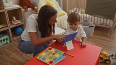 Anne ve küçük oğlu, oyuncak ve kitaplarla rahat çocuk odasında birlikte tablet keşfedip kapalı alanda etkileşimli bir öğrenme ortamı yaratıyorlar..