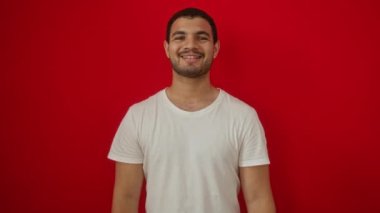 Beyaz tişört giyen genç İspanyol adam ayakta ve gülümserken mutlu bir şekilde işaret dilini gözünün üstüne koy, parmaklarını parmaklarının arasından geçir, kırmızı arka planda izole edilmiş.