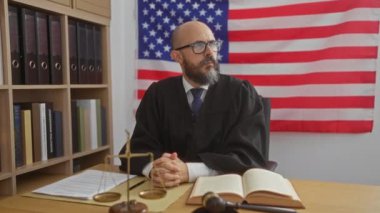 Yargıç cüppeli bir adam hukuk kitapları, tokmak ve tartı ile bir mahkeme salonunda arka planda Amerikan bayrağı ile bir masada oturuyor.
