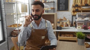 Fırında çalışan sakallı genç adam akıllı telefonla konuşurken tablet kullanıyor.