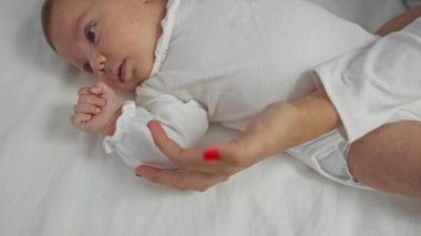 Bebek yatakta uzanmış bir kadınla nazikçe el ele tutuşuyor, kapalı bir aile sahnesinde ilişki, sevgi ve bakım sergiliyor..