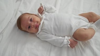 Beyaz elbiseli yeni doğmuş erkek bebek, sıcak yatak odası ortamında sevimli bir şekilde yatakta uzanıyor, çocukluk masumiyetinin ve sıcaklığın özünü yakalıyor.